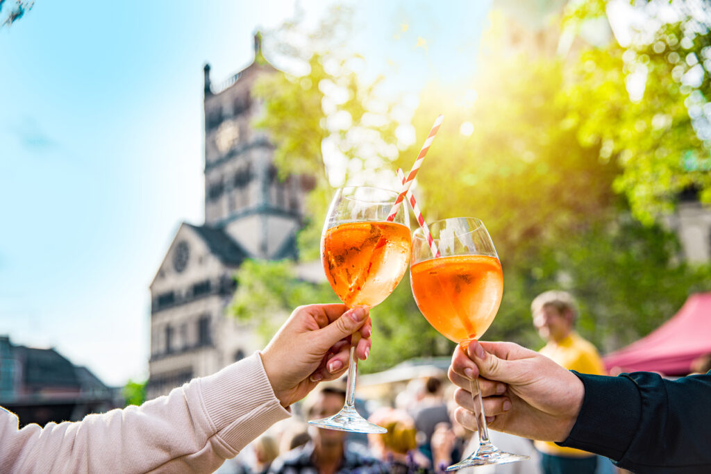 Zwei Personen stoßen mit einem Glas Aperol auf dem Feierabendmarkt an. Im Hintergrund sieht man den Turm des Quirinus-Münsters