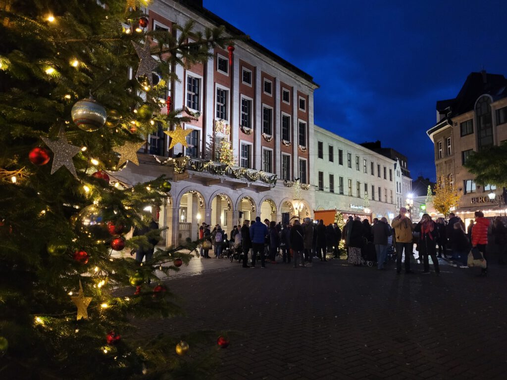 Vorne stet ein geschmückter Tannenbaum. Im Hintergrund sind mann einige Menschen sowie das Rathaus im Lichterglanz