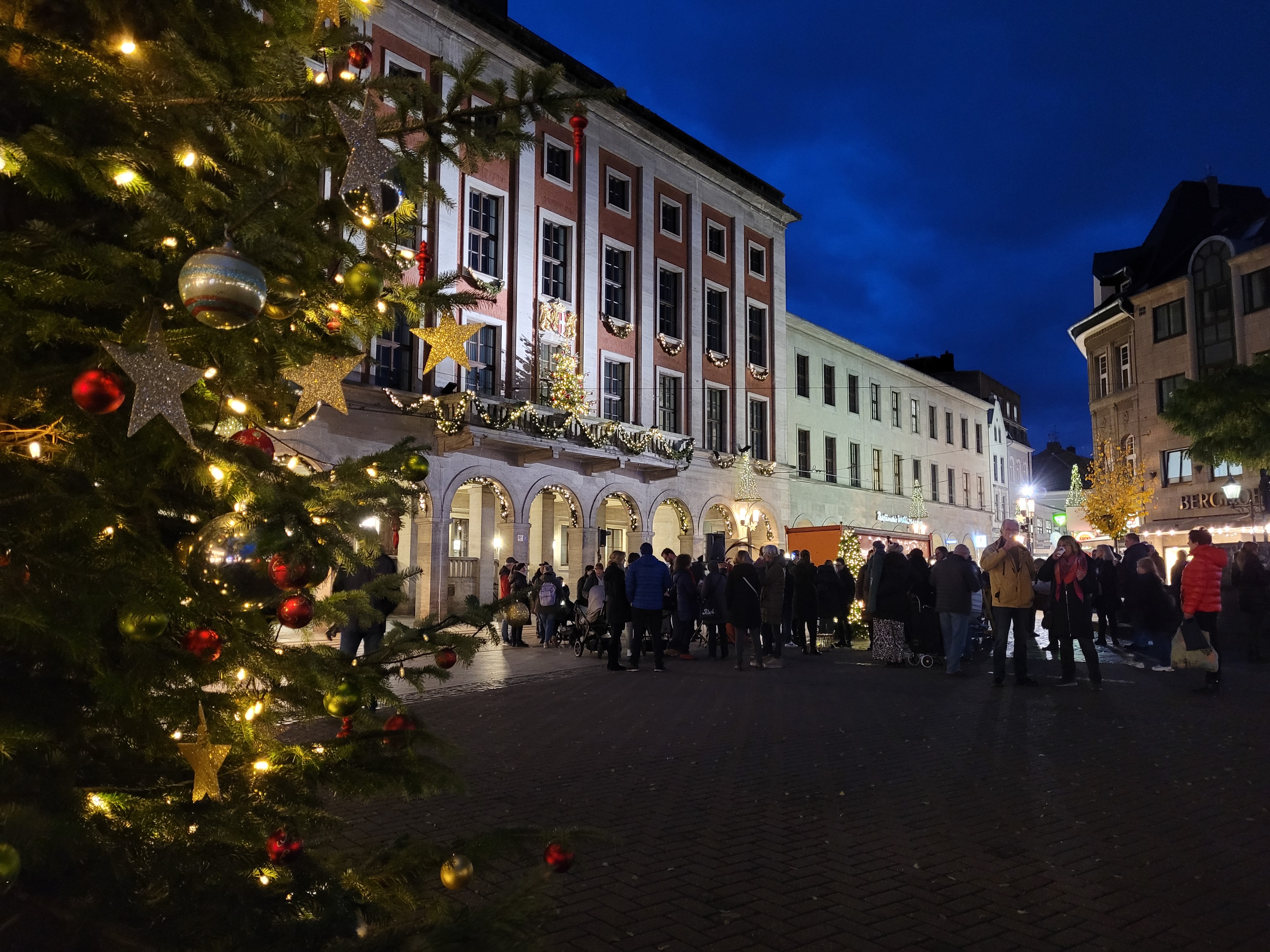 Vorne stet ein geschmückter Tannenbaum. Im Hintergrund sind mann einige Menschen sowie das Rathaus im Lichterglanz
